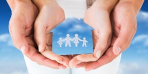 Close up of hands gently cupping a small blue paper house with a cut out family silhouette inside, set against a soft sky background, symbolising Mortgage Life Insurance UK